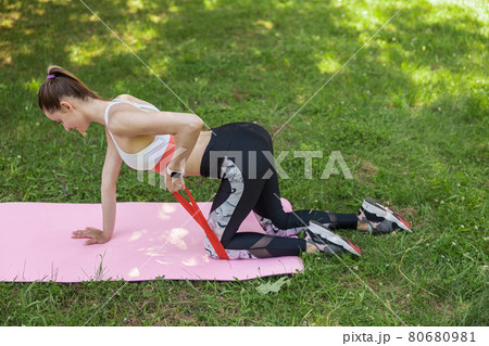 Attractive lady in sportswear does exercises for arms with elastic band kneeling on green lawn 80680981