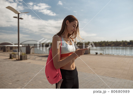 Positive woman in stylish tracksuit uses mobile phone standing on city waterfront 80680989