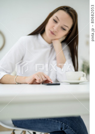 Young brunette in a white shirt is waiting for a business partner in a cafe. Woman at a business meeting. Girl drinks coffee during the break and uses a smartphone reads sms. Young brunette in a white shirt is waiting for a business partner in a cafe. Woman at a business meeting. Girl drinks coffee during the break and uses a smartphone reads sms. 80682053