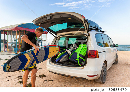 Back view young adult male surfer in wetsuit put out surf board kite equipment on sand beach from van vehicle with rooftop box ocean sea beach watersport spot camp.Adventure travel sport acitivity 80683479