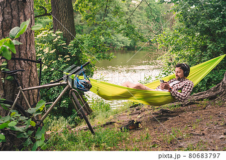 Outdoors and adventure concept. Bike trip to forest. Cyclist is resting in green hammock between trees in nature by lake while listening to music on headphones and surfing Internet on smartphone 80683797