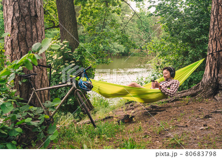 Young caucasian man resting in hammock, listening to music on headphones and using smartphone afterwards on bicycle in forest near the lake. Audio healing. Idyllic place. Travel, camping in nature 80683798
