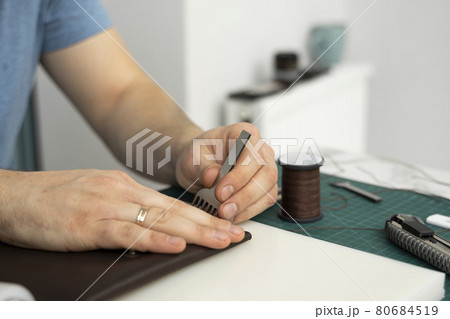Mens hand holding a leather carving hammer and steel chisel hole punch and makes a leather wallet in his workshop. Working process with a brown natural leather. Craftsman holding a crafting tools. Mens hand holding a leather carving hammer and steel chisel hole punch and makes a leather wallet in his workshop. Working process with a brown natural leather. Craftsman holding a crafting tools. 80684519