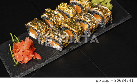 Custom sushi roll with nori, cuttlefish ink, fresh salmon, cream cheese, pepper pumpkin, eel, unagi sauce and edible gold leaf with red ginger, wasabi on a square plate on black table and background. Custom sushi roll with nori, cuttlefish ink, fresh salmon, cream cheese, pepper pumpkin, eel, unagi sauce and edible gold leaf with red ginger, wasabi on a square plate on black table and background. 80684541