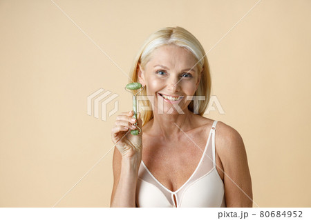 Portrait of mature blonde woman with perfect glowing skin smiling at camera, holding jade facial roller for skin care and beauty treatment, posing isolated over beige background 80684952