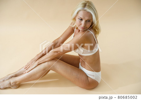 High angle view of mature blonde woman with perfect body posing in underwear, sitting on studio floor and looking away isolated over beige background 80685002