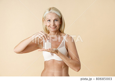 Adorable mature woman in underwear holding and opening white jar of skin care cream or lotion, standing isolated over beige background Adorable mature woman in underwear holding and opening white jar of skin care cream or lotion, standing isolated over beige background 80685003