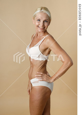 Pretty mature woman in underwear with fit body turning head to camera and smiling, holding hand on waist standing isolated over beige background 80685449