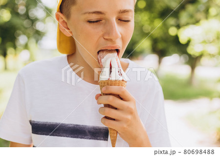 Teenager boy eating ice-cream cone on green nature background. Summer, junk food and people concept Teenager boy eating ice-cream cone on green nature background. Summer, junk food and people concept 80698488
