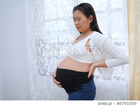 Young beautiful pregnant woman suffering from pain in the room. touching her belly and keeping one hand on her back.	 80701030