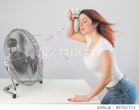 A red-haired Caucasian woman chills by the electric fan and drinks a cold drink. Climate control in the apartment 80701711