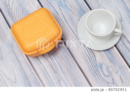 Plastic yellow lunch box and cup with saucer on wooden background. Plastic yellow lunch box and cup with saucer on wooden background. 80702951