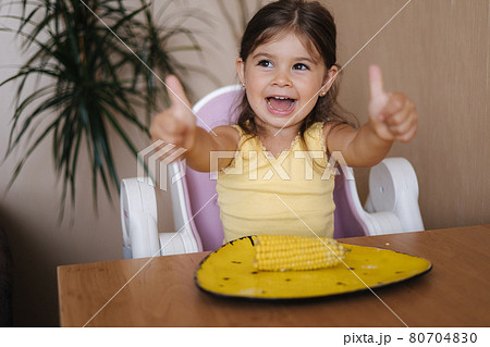 Happy little girl hold fresh corn in hand and smile. Female kid thumbs up. Summer mood at home Happy little girl hold fresh corn in hand and smile. Female kid thumbs up. Summer mood at home 80704830