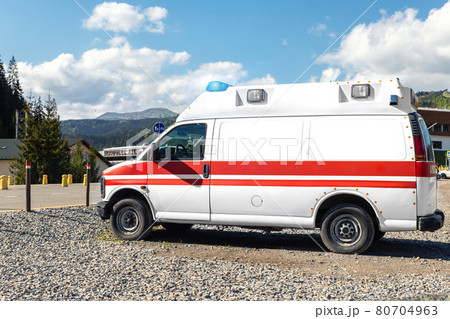 Side view of white ambulance rescue ems van car parked near countryside rural road at highland mountain resort area. Paramedic first aid help service vehicle against alpine forest landscape 80704963