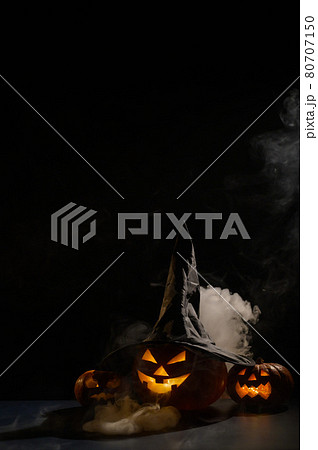 Vertical Halloween card. Witch hat on a pumpkin with carved creepy grimaces on a black background in the fog. Jack-o-lantern glows from the inside and emits smoke. Vertical Halloween card. Witch hat on a pumpkin with carved creepy grimaces on a black background in the fog. Jack-o-lantern glows from the inside and emits smoke. 80707150