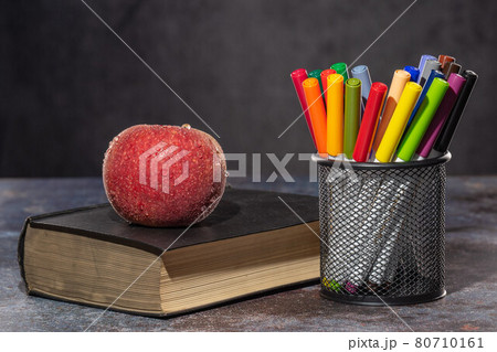 Back to school. Apple in drops of water lying on the book, next to multicolored markers standing in the stand Back to school. Apple in drops of water lying on the book, next to multicolored markers standing in the stand 80710161