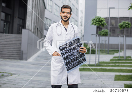 Male Arab doctor smiling at camera, holding tomography scan, standing outdoors near clinic. 80710673