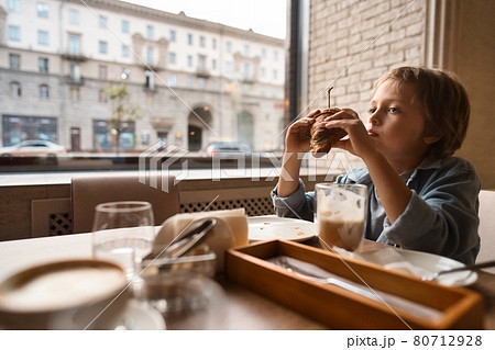 Happy cute boy eating fresh croissant, sitting at the table in city cafe, with paper cup of cocoa Happy cute boy eating fresh croissant, sitting at the table in city cafe, with paper cup of cocoa 80712928