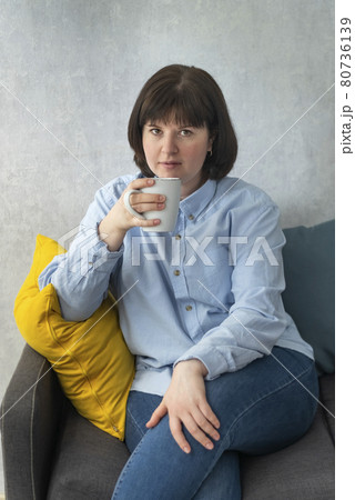 Woman is holding mug of coffee and sitting on sofa in living room. Coffee break. Vertical frame Woman is holding mug of coffee and sitting on sofa in living room. Coffee break. Vertical frame 80736139