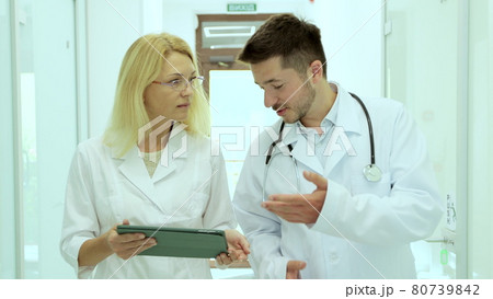 Team of physicians wearing lab coat discussing patient medical test walking in hospital corridor. Diverse doctors studying diagnosis on clipboard in clinic hallway. 80739842