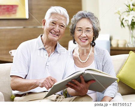 senior asian couple sitting on couch holding a book looking at camera smiling senior asian couple sitting on couch holding a book looking at camera smiling 80750517