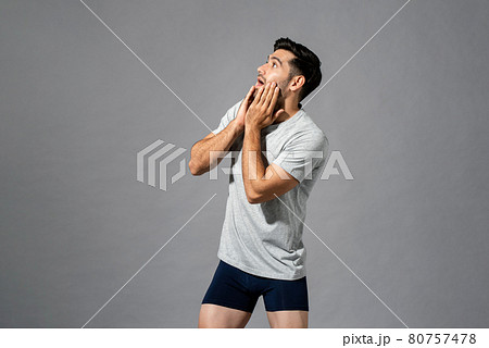 Surprised young Caucasian man in sleepwear with hands on cheeks looking up to empty space aside 80757478