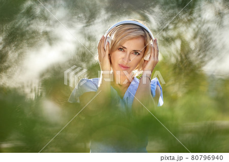 Blond woman in wireless headphones in public park. Blond woman in wireless headphones in public park. 80796940