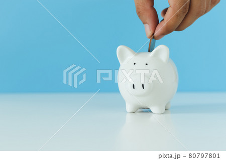 White piggy bank with hand putting money coins on blue background, copy space. White piggy bank with hand putting money coins on blue background, copy space. 80797801