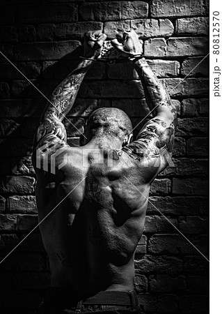 A muscular male prisoner stands with his hands up in handcuffs facing the wall. 80812570