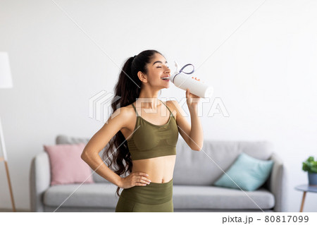 Pretty Indian woman in sports clothes drinking water or protein shake from bottle at home 80817099