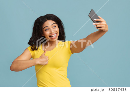 Excited african american lady taking selfie on smartphone and showing thumb up over blue background Excited african american lady taking selfie on smartphone and showing thumb up over blue background 80817433