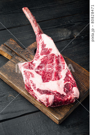 Dry aged raw tomahawk beef steak with ingredients for grilling, on black wooden table background Dry aged raw tomahawk beef steak with ingredients for grilling, on black wooden table background 80828471