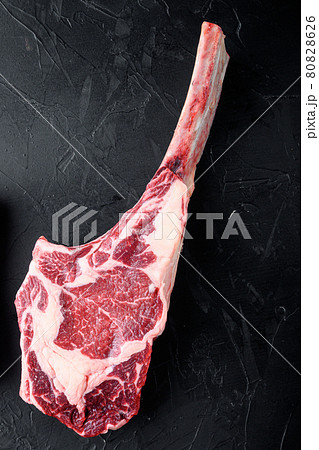 Dry aged raw tomahawk beef steak with ingredients for grilling, on black stone background, top view flat lay, with copy space for text 80828626