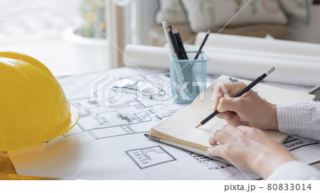 Architectural building design and construction plans with blueprints, young man is taking notes and planning a house or building with eraser, pen, pencil, tape measure, architect hat other equipment. Architectural building design and construction plans with blueprints, young man is taking notes and planning a house or building with eraser, pen, pencil, tape measure, architect hat other equipment. 80833014