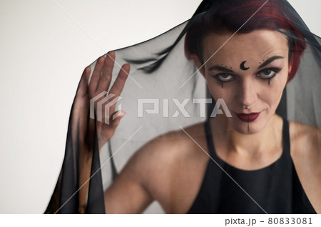 Redhead woman with a black veil on her head in a dark studio. A girl with an earring in her nose smiles slyly. Witch Makeup. Halloween costume. 80833081