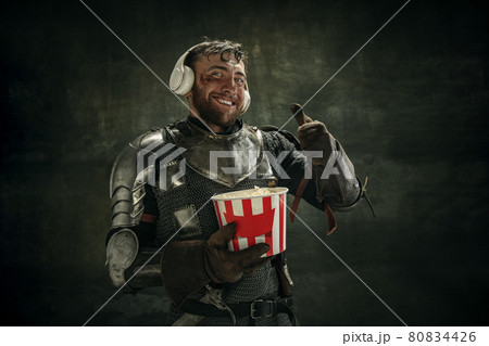 Portrait of one brutal bearded man, medeival warrior or knight in headphones with bucket of popcorn over dark background. Portrait of one brutal bearded man, medeival warrior or knight in headphones with bucket of popcorn over dark background. 80834426