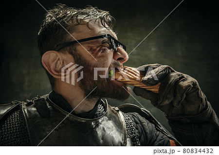 Portrait of one brutal bearded man, medeival warrior or knight with hot dog and cool drink over dark background. 80834432