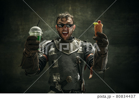 Portrait of one brutal bearded man, medeival warrior or knight with hot dog and cool drink over dark background. 80834437