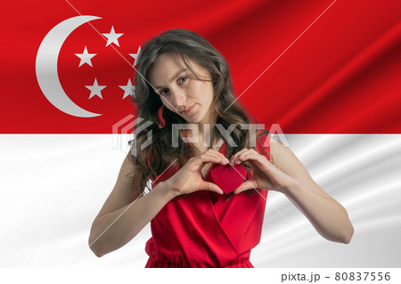 Love Singapore. A girl holds a heart on her chest in her hands against the background of the flag of Singapore. The concept of patriotism 80837556