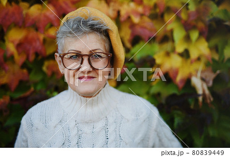 Senior woman standing outdoors against colorful natural autumn background, looking at camera. 80839449