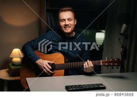Medium shot of cheerful guitarist singer man emotionally singing into condenser microphone and playing acoustic guitar in dark home recording studio. 80843668
