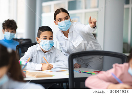 Female teacher in surgical face mask helping small hispanic schoolboy Female teacher in surgical face mask helping small hispanic schoolboy 80844938