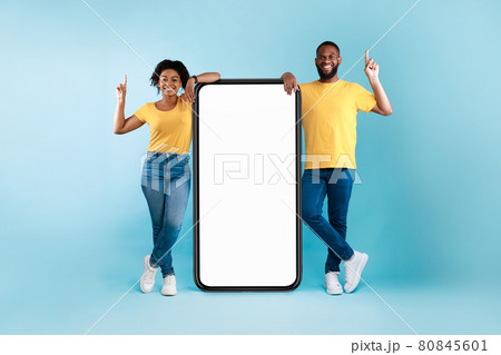 Cheerful Afro couple standing next to big smarthone with blank screen, pointing upwards, recommending mobile app, mockup Cheerful Afro couple standing next to big smarthone with blank screen, pointing upwards, recommending mobile app, mockup 80845601