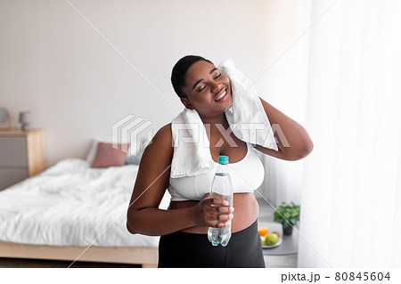 Plus size black woman with bottle of water wiping sweat with towel after domestic workout, indoors 80845604