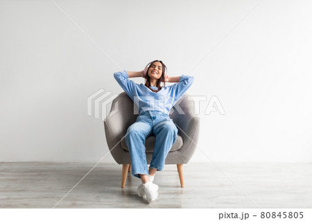Full length of young Caucasian woman relaxing in armchair with hands behind her head, listening to music in headphones 80845805