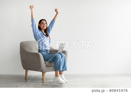Ecstatic woman sitting in armchair with laptop, lifting hands up, making YES gesture, celebrating online win or success 80845976
