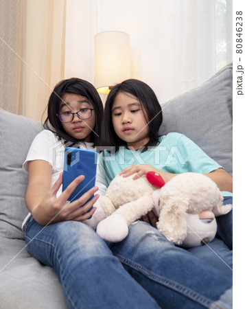 Happy siblings children sitting on sofa in living room speak with parent on mobile phone together, smiling elder girl showing cute little sister video call by smartphone speak with family. Happy siblings children sitting on sofa in living room speak with parent on mobile phone together, smiling elder girl showing cute little sister video call by smartphone speak with family. 80846238