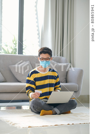 Young male shopper in medical face mask using laptop on couch for online shopping 80865941