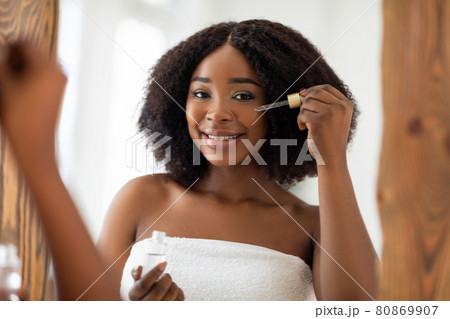Smiling young black lady in towel using facial serum near mirror, taking care about her beauty at home Smiling young black lady in towel using facial serum near mirror, taking care about her beauty at home 80869907