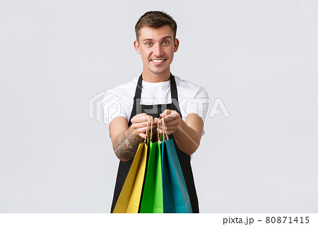 Retail store, shopping and employees concept. Friendly handsome and polite salesman handing over eco-bags with purchased items to client in shop, standing white background 80871415
